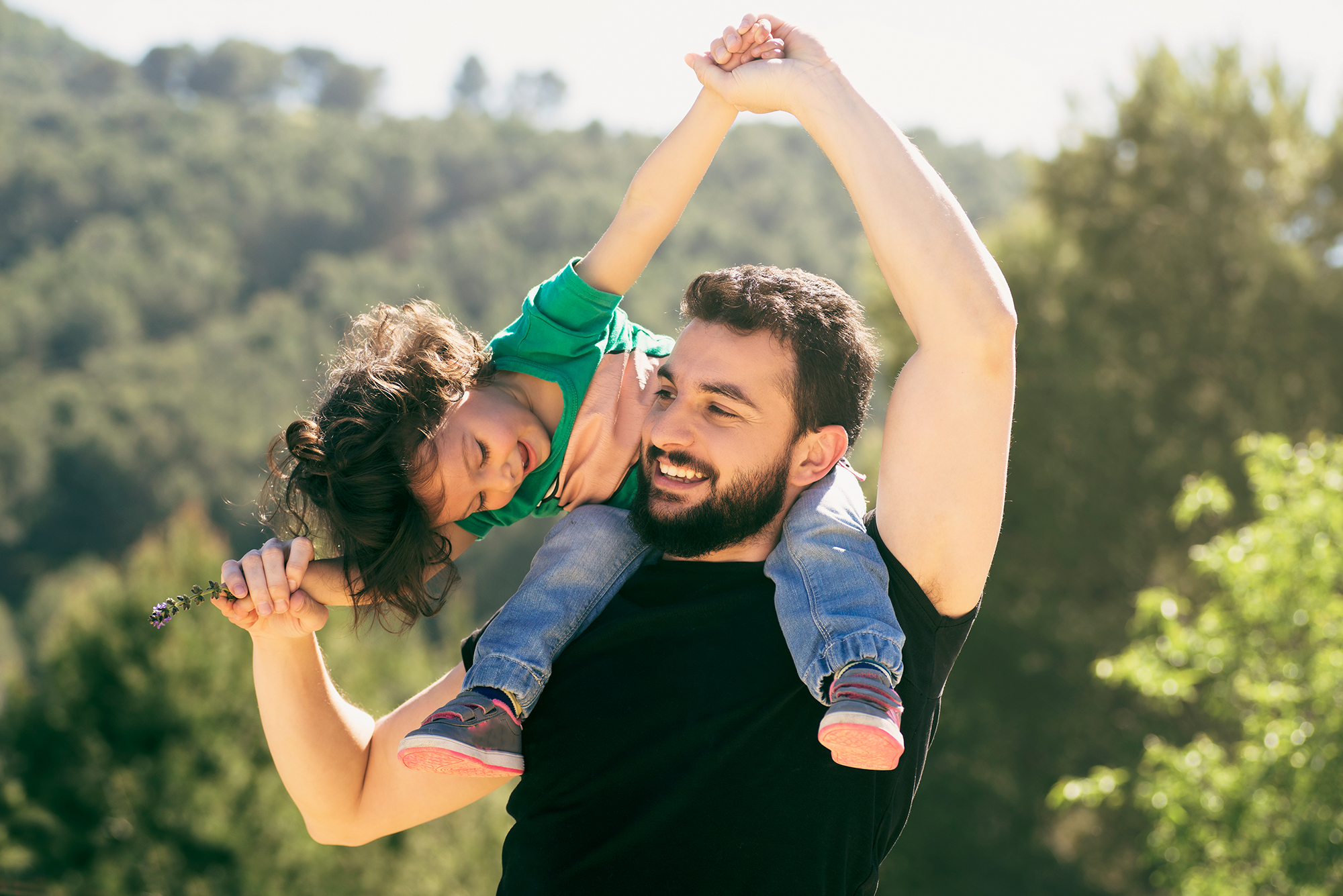 A daughter is sitting on her fathers shoulders happily playing outdoors