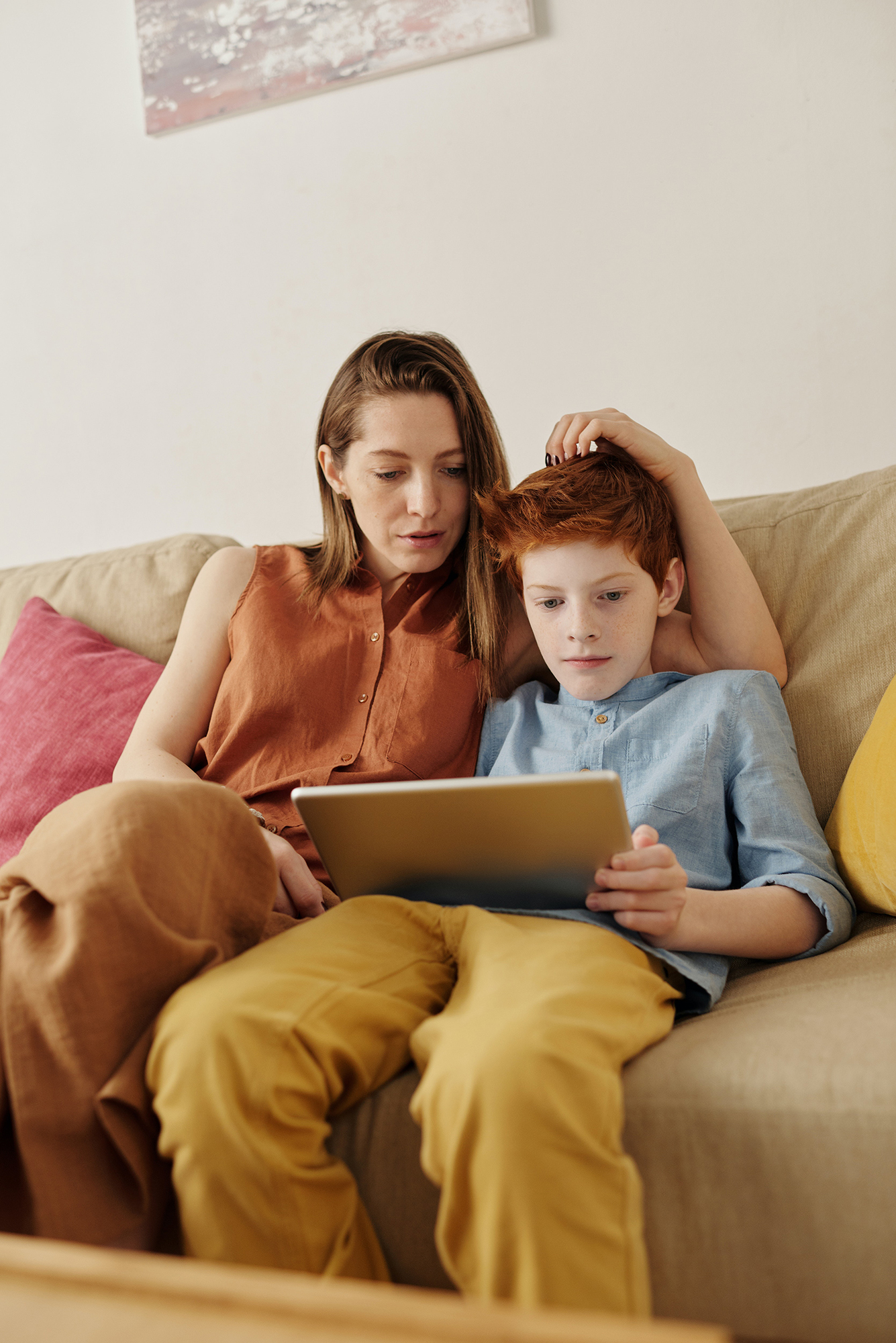 A mother and son sit on a couch and both look at the screen of a tablet